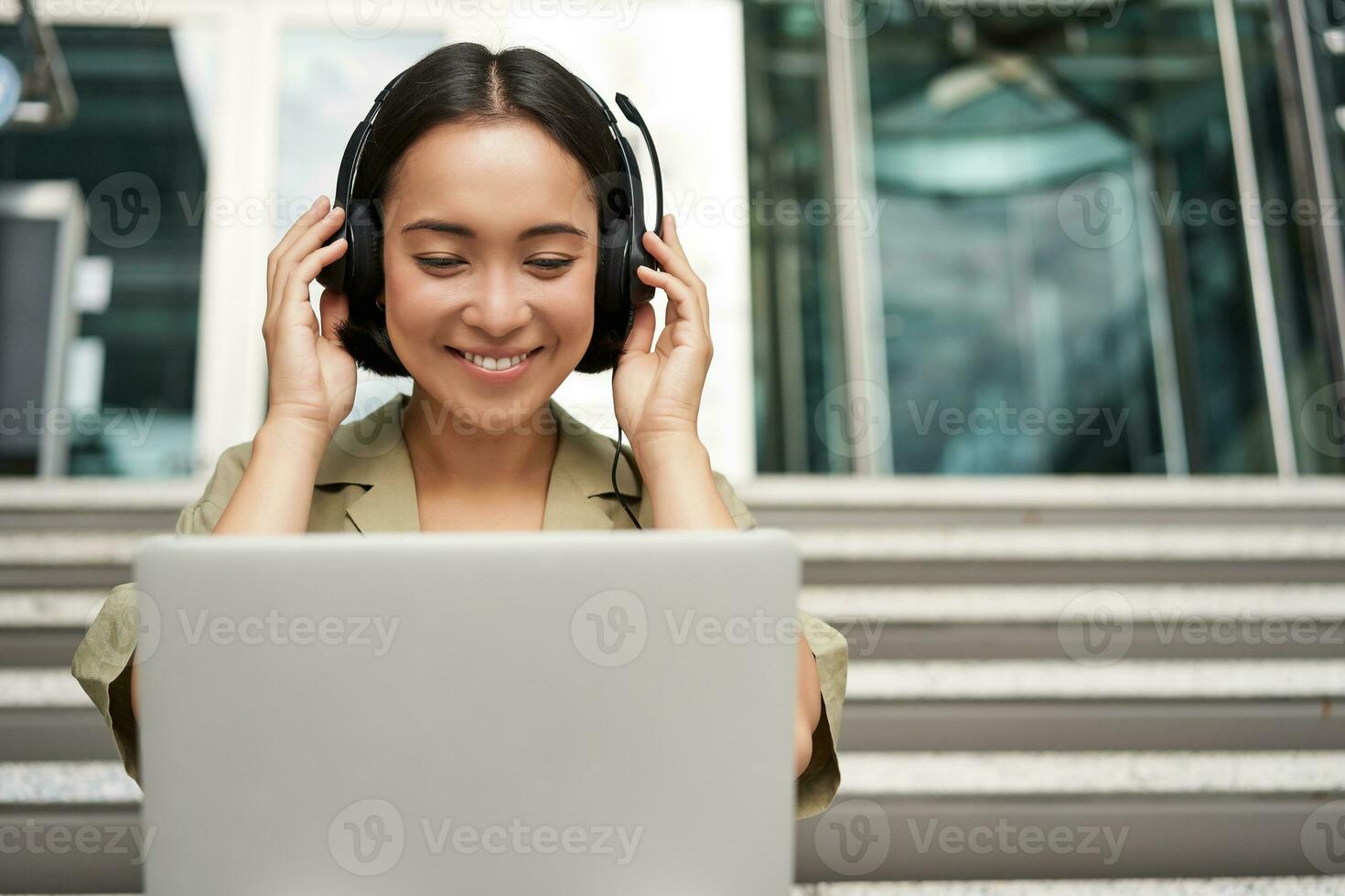 Portrait of young asian woman sitting with laptop and headphones, watching video, does online course on computer, sitting on stairs outdoors photo