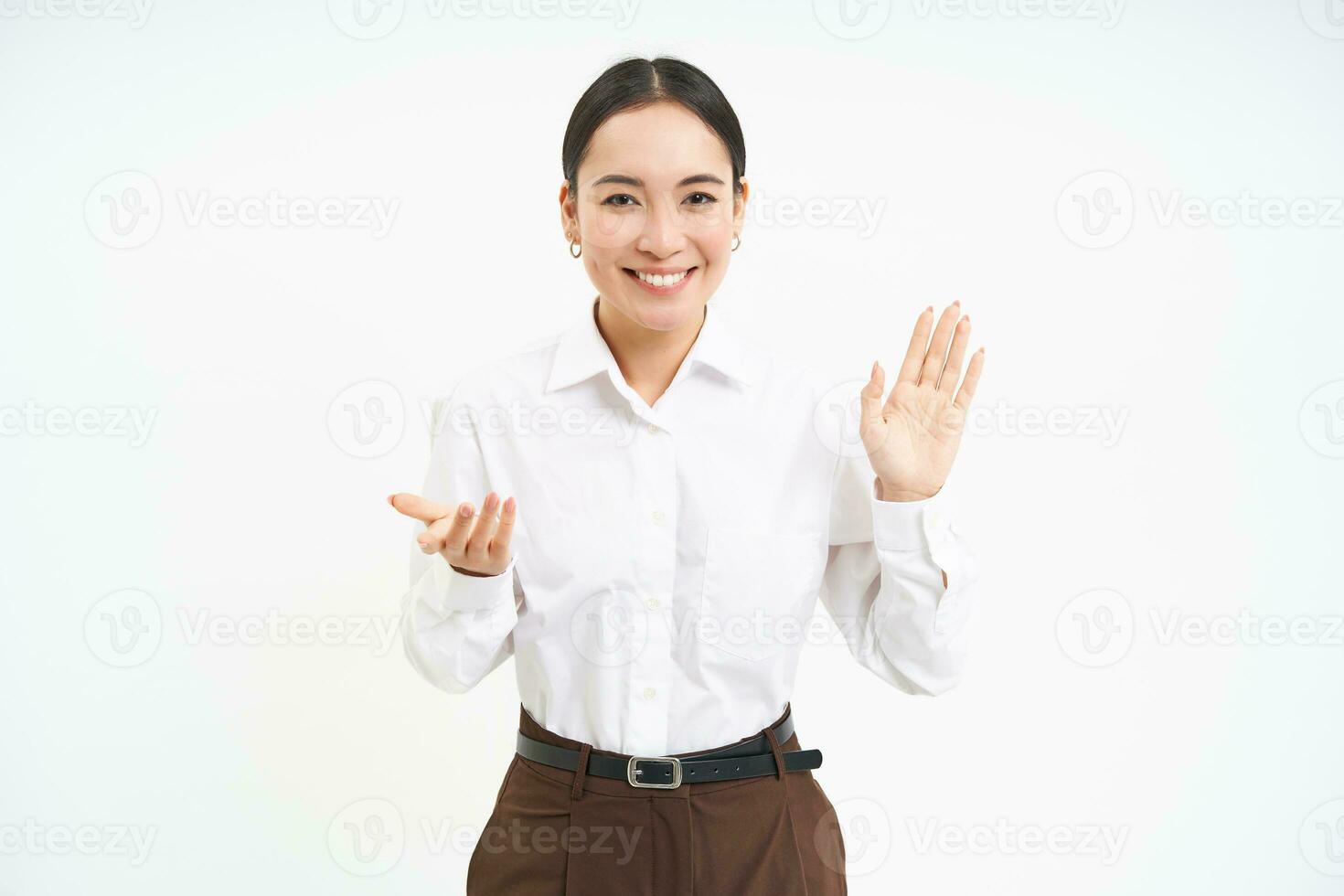 Portrait of beautiful asian woman raises one hand, smiles friendly, introduces herself, greets and says hello, white background photo