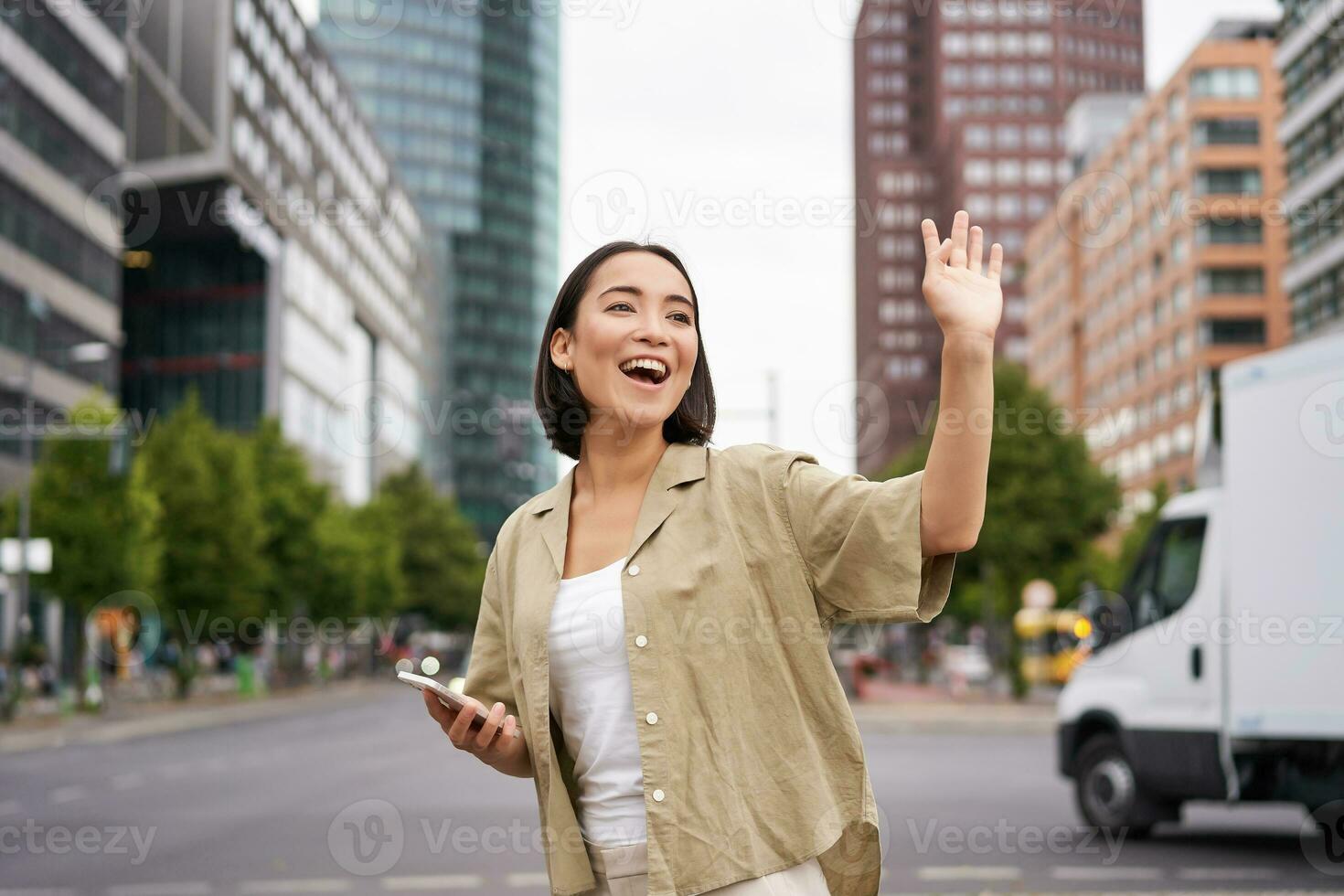 Happy asian girl passing by friend and waving at them on street, saying hello while walking in city, holding smartphone photo