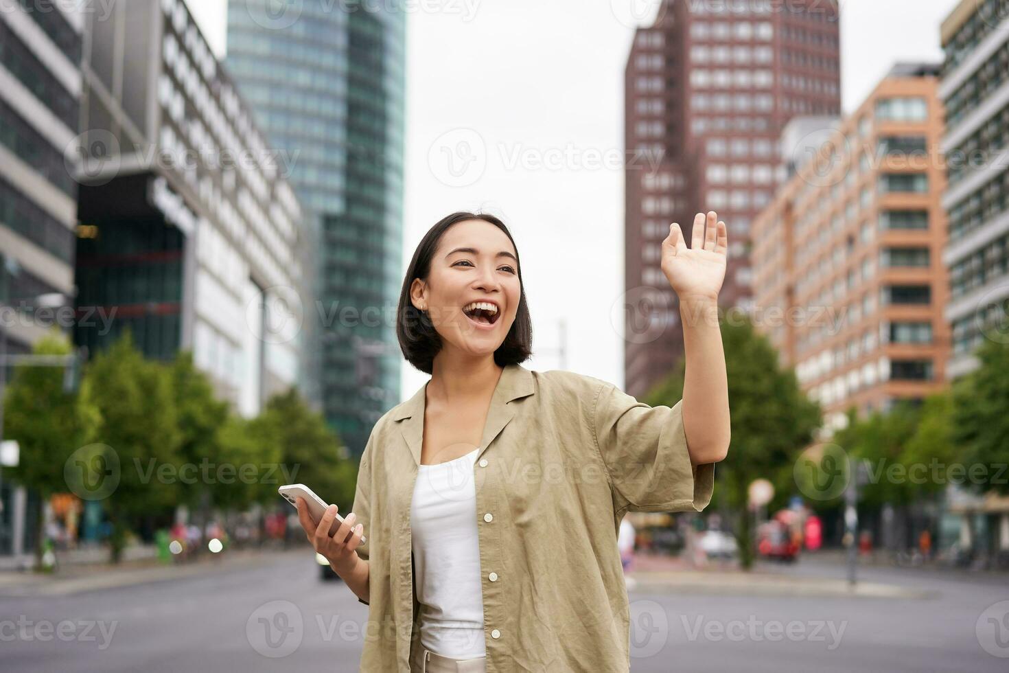 Happy asian girl passing by friend and waving at them on street, saying hello while walking in city, holding smartphone photo