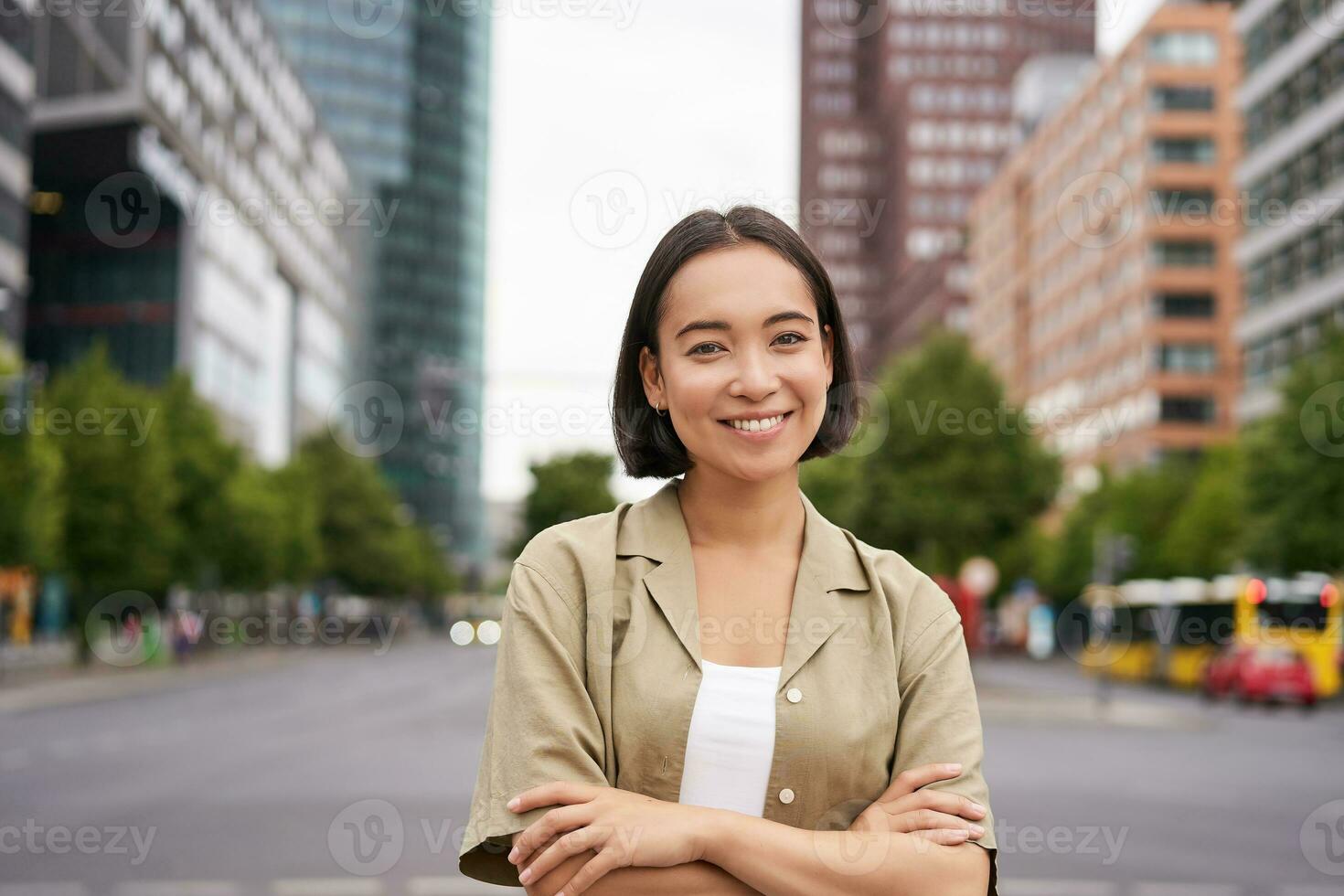 Urban people. Young happy asian girl cross arms on chest, posing on busy city street, smiling with confidence at camera photo