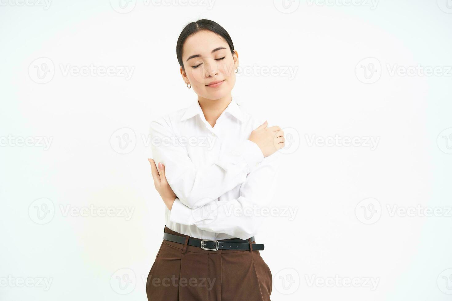 Self-hug and wellbeing. Young businesswoman smiles, embraces herself, wraps hands around her body with happy face, white background photo