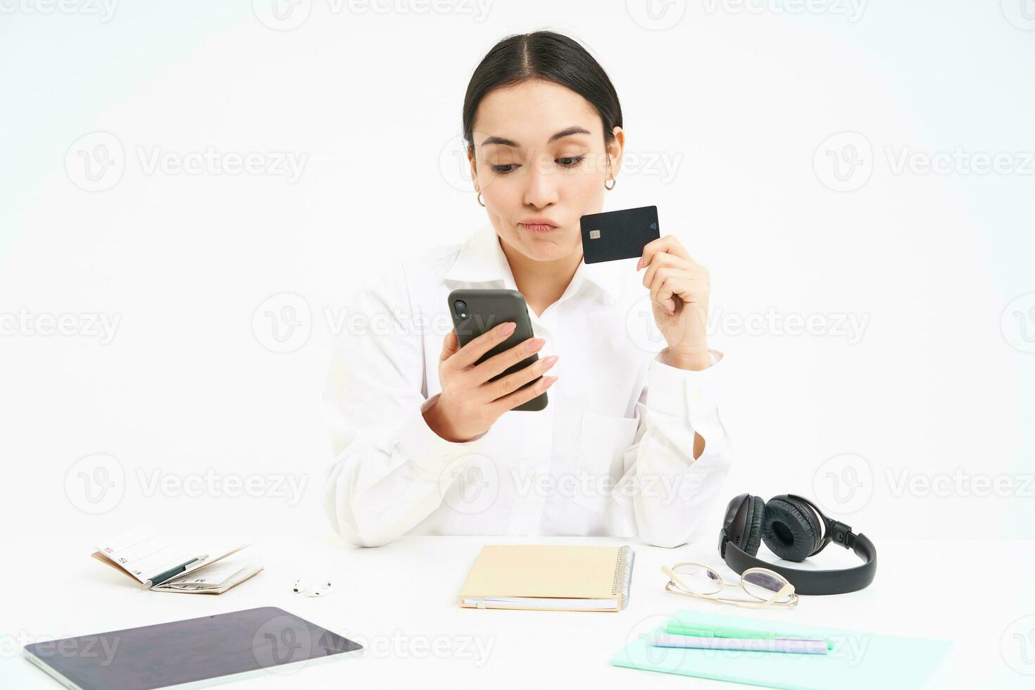 Young woman student, sits with smartphone and credit card, looks shocked at her bank account on mobile phone app, sitting frustrated over white background photo