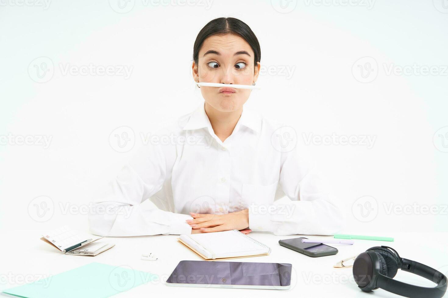 negocio y oficina trabajar. joven mujer sentado a lugar de trabajo, engañando alrededor, sostiene bolígrafo con labios y muestra gracioso caras, blanco antecedentes foto