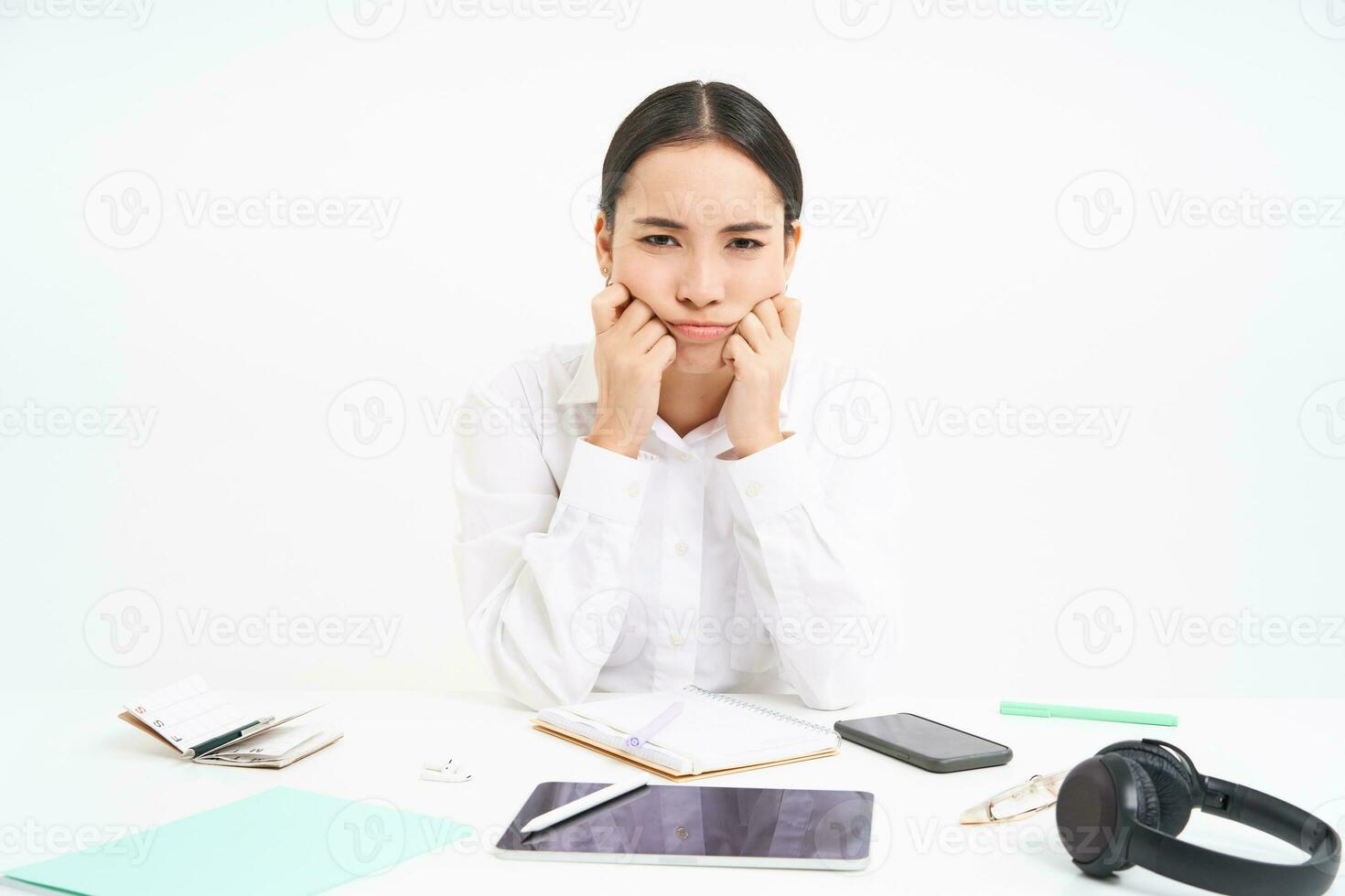 Sad and grumpy asian woman at workplace, sits with digital tablet and work documents, looks upset and bored, white background photo