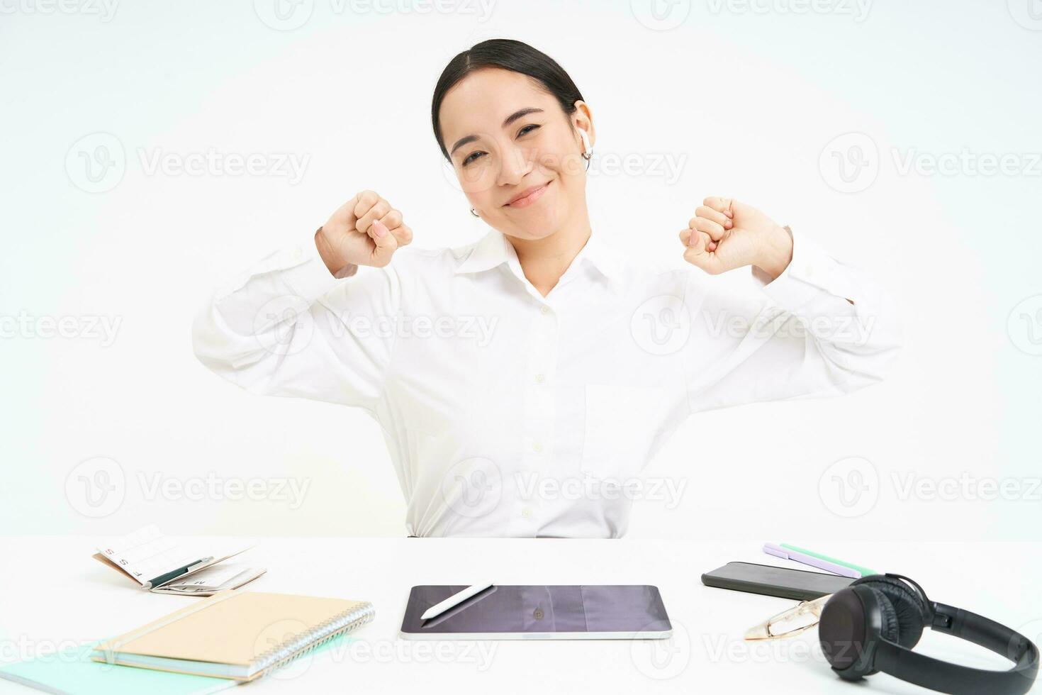 Workplace and business concept. Smiling beautiful asian woman, office worker sits in office, stretches her hands with pleased face after completed work, white background photo