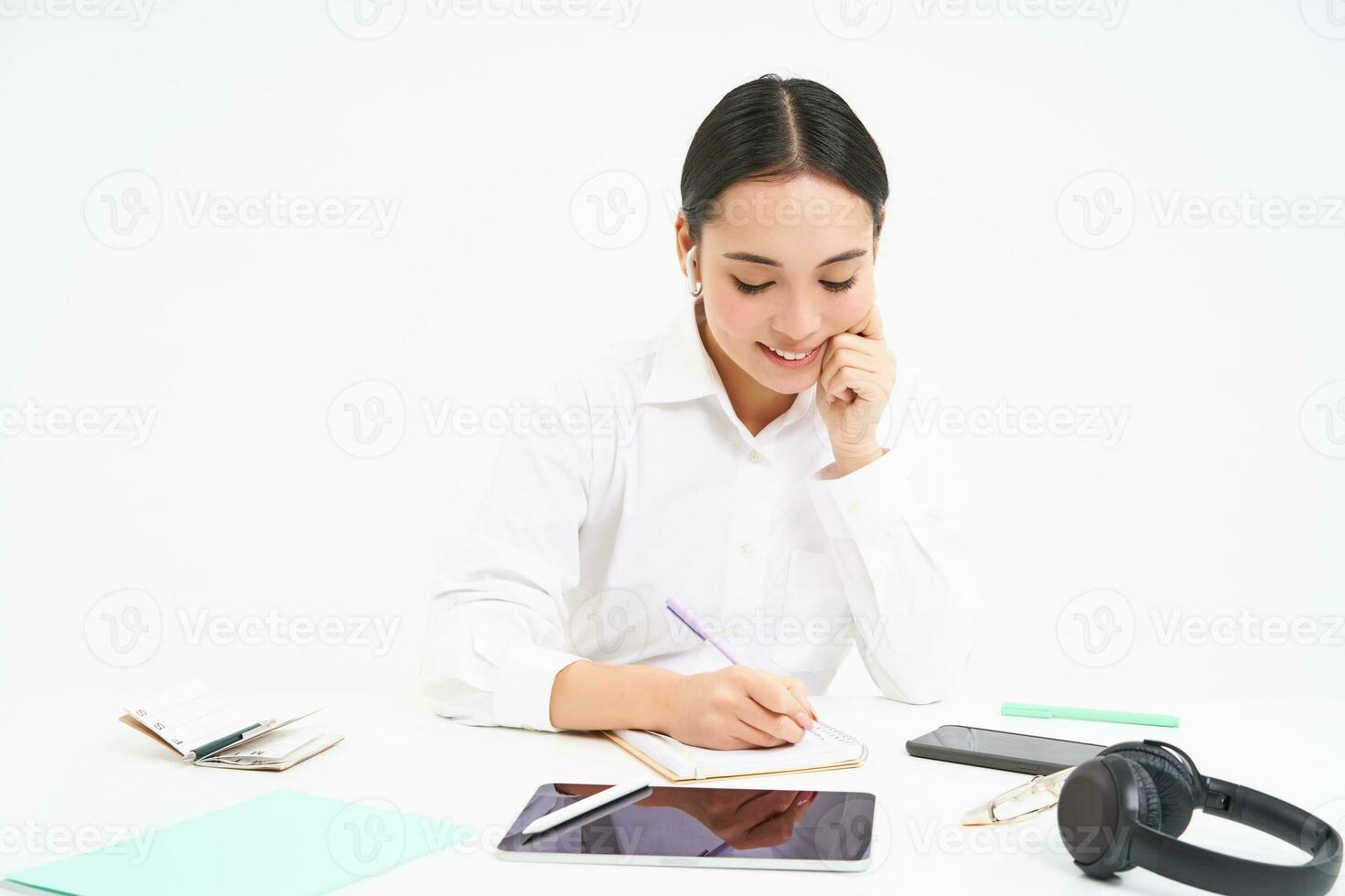 Asian working woman sits in her office, looks at tablet and writes down information, isolated on white background photo