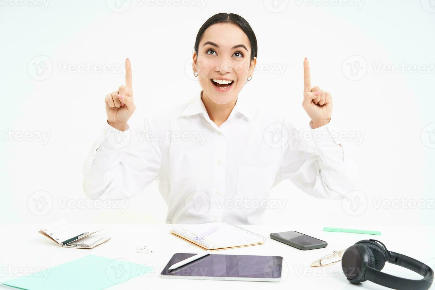 Business and profession. Portrait of asian woman in workplace, sits in office and points up, shows banner advertisement, white background photo