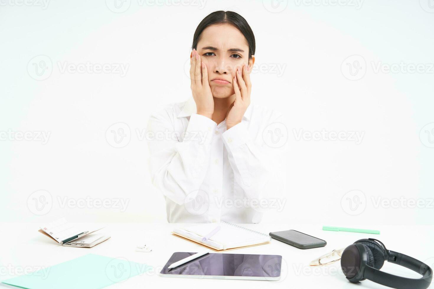 Sad and grumpy asian woman at workplace, sits with digital tablet and work documents, looks upset and bored, white background photo