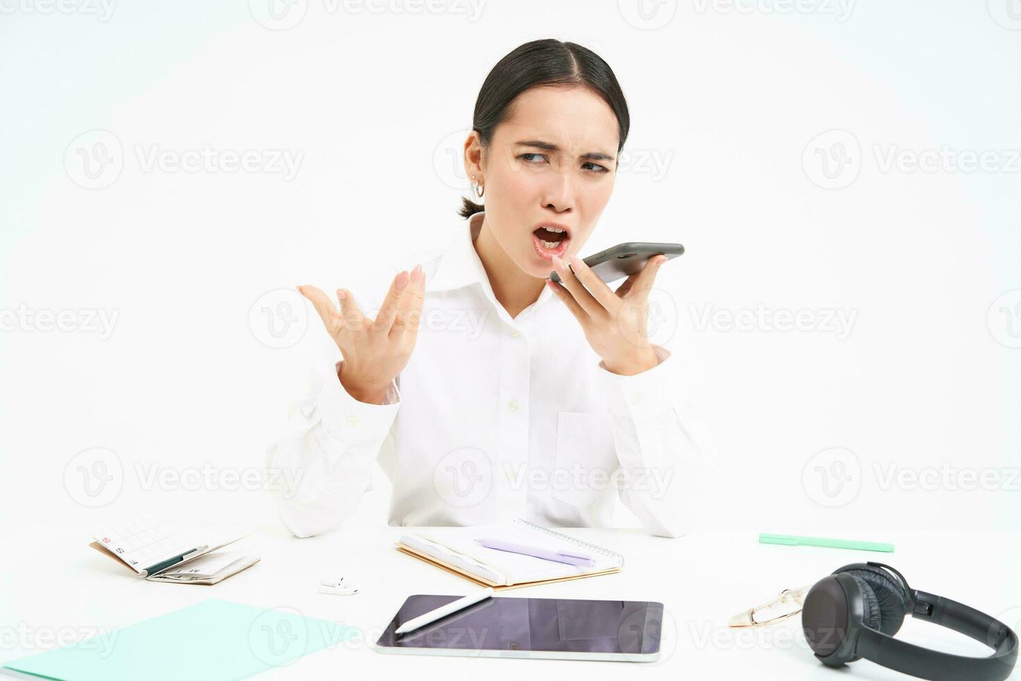 Portrait of angry korean employer, lady boss sits in office with mobile phone, shouts with annoyed face at cellphone, has argument, white background photo