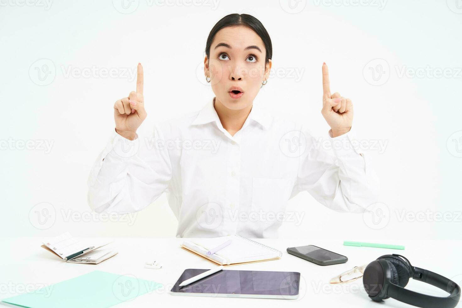 Business and profession. Portrait of asian woman in workplace, sits in office and points up, shows banner advertisement, white background photo