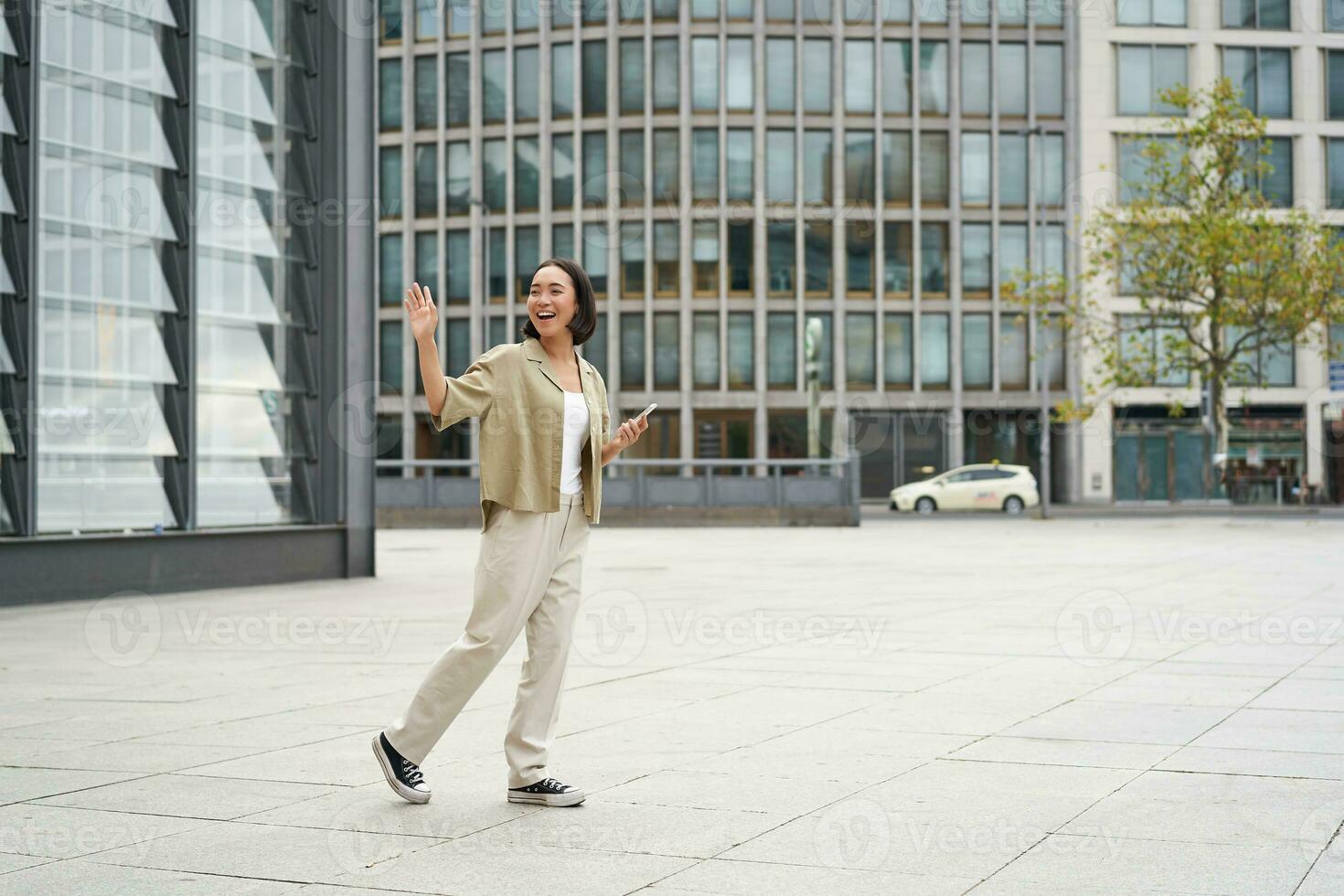 Happy asian girl saying bye to someone, waving at friend and walking on street with smartphone photo