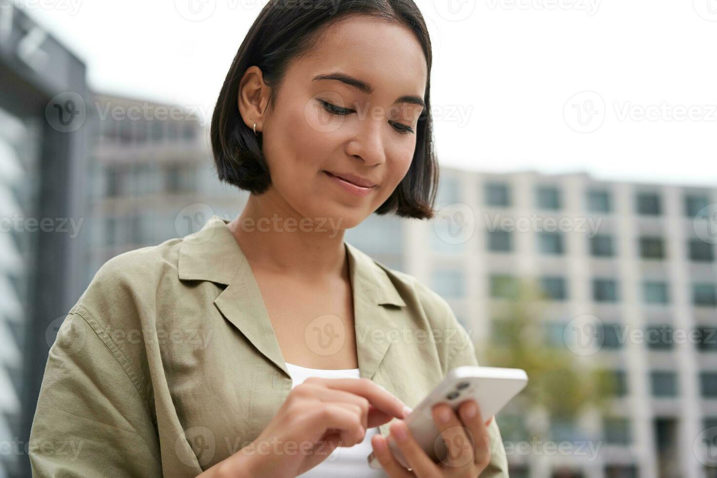 Mobile technology. Smiling asian woman using smartphone app, looking at her telephone on street, checking map, calling or texting someone photo