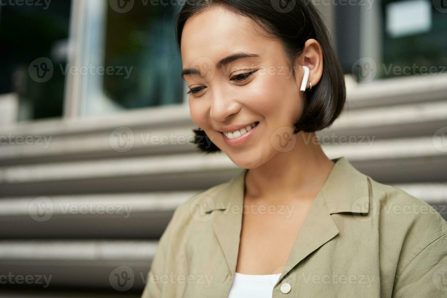 retrato de sonriente asiático niña escucha música, podast en inalámbrico auriculares, utilizando auriculares al aire libre, sentado en calle foto