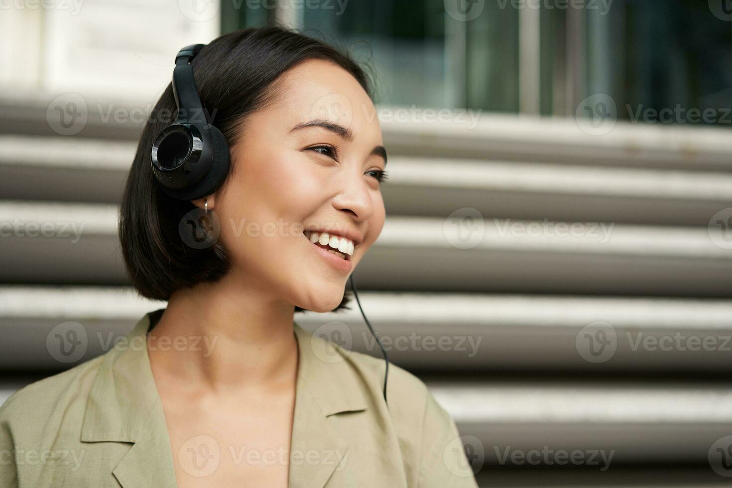 sonriente asiático chica, reír, escuchando música en auriculares, sentado al aire libre. uni estudiante disfrutando gratis hora foto