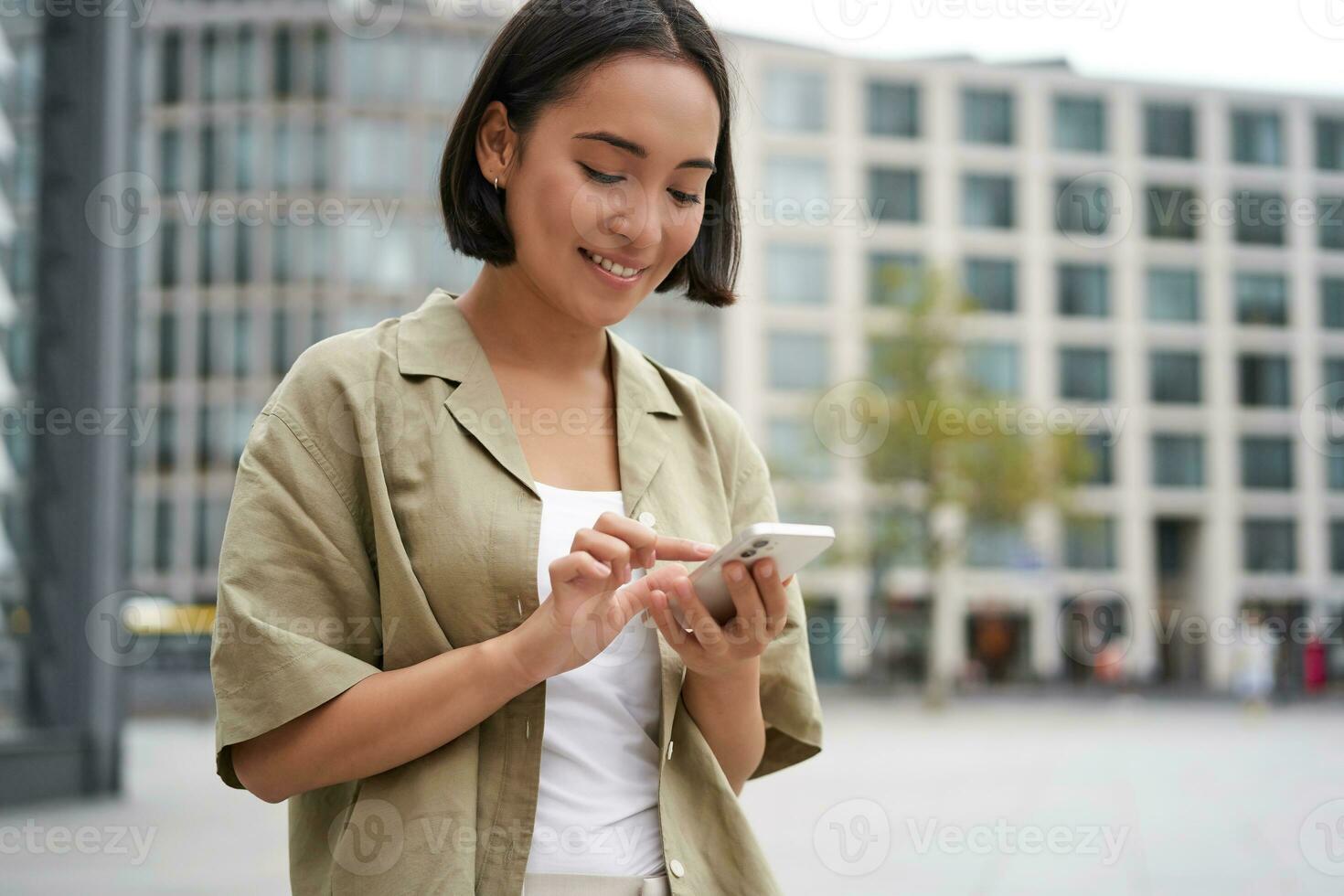 Mobile technology. Smiling asian woman using smartphone app, looking at her telephone on street, checking map, calling or texting someone photo