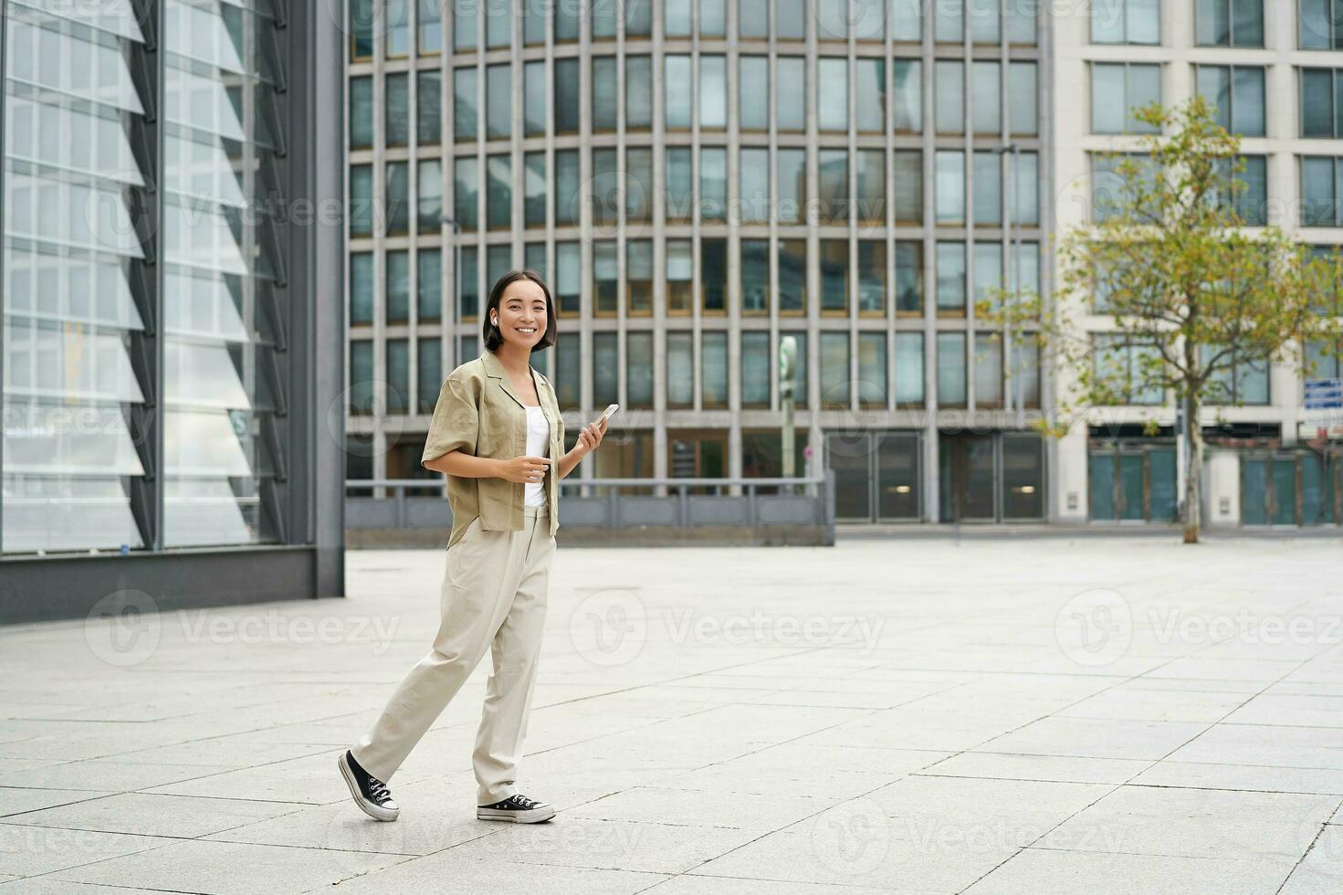 Portrait of beautiful smiling asian woman, walking on street of city centre with smartphone, looking at camera photo