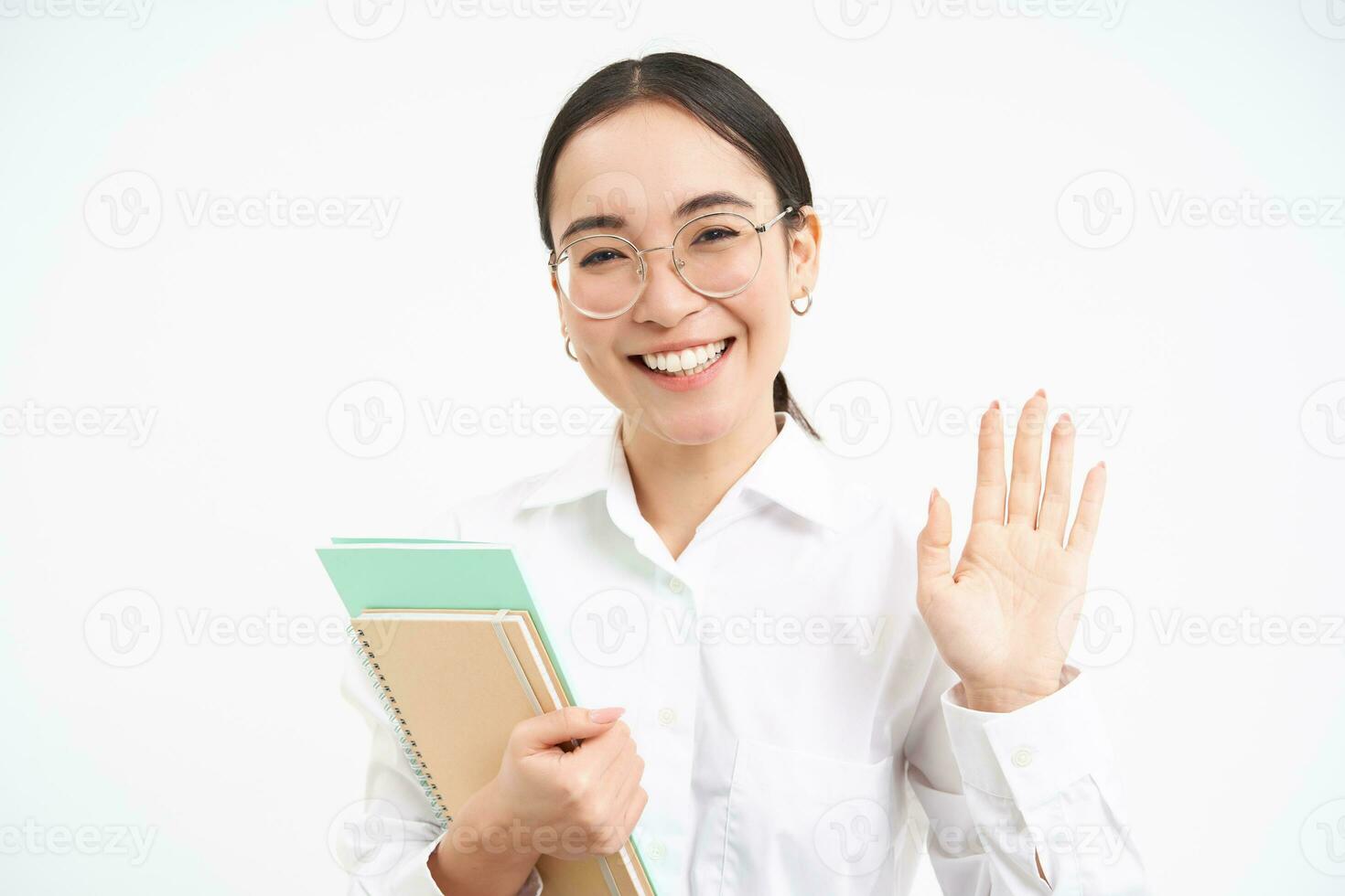 Image of friendly japanese student, girl waves hand, says hello, wears glasses, holds notebooks, stands over white background photo