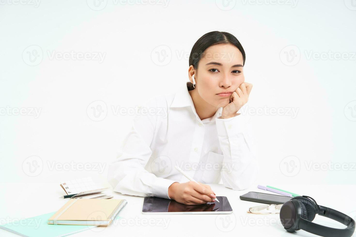 cansado y aburrido coreano mujer en oficina, empleado se sienta con digital tableta, escucha música en auriculares y mira cansado, blanco antecedentes foto