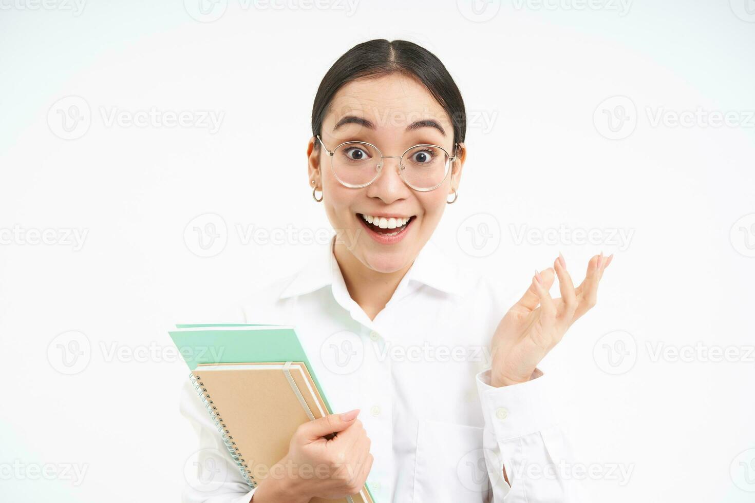Portrait of corporate woman, asian woman in glasses, holds notebook and looks surprised, amazed by smth, stands over white background photo