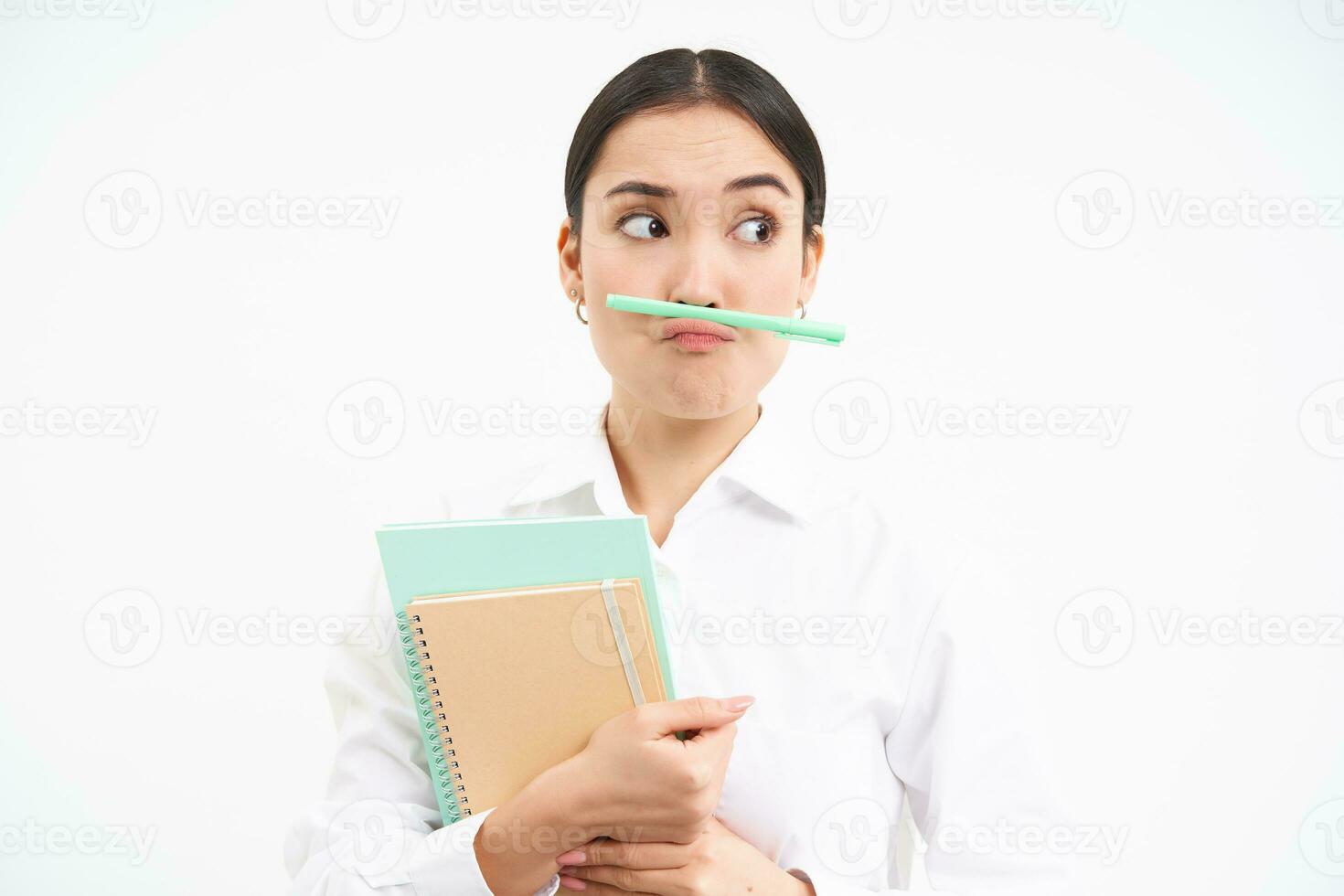 gracioso coreano mujer con cuadernos, sostiene bolígrafo con labios y nariz y hace caras, soportes terminado blanco estudio antecedentes foto