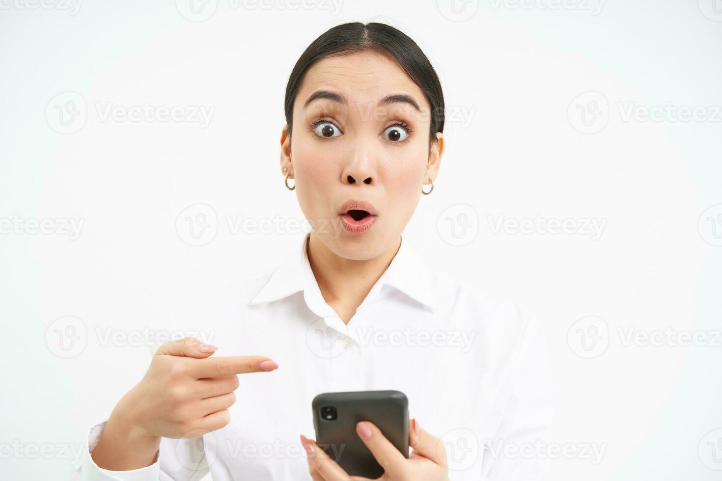 Close up portrait of happy saleswoman, successful woman with smartphone, pointing at her phone and smiling amazed, standing over white background photo
