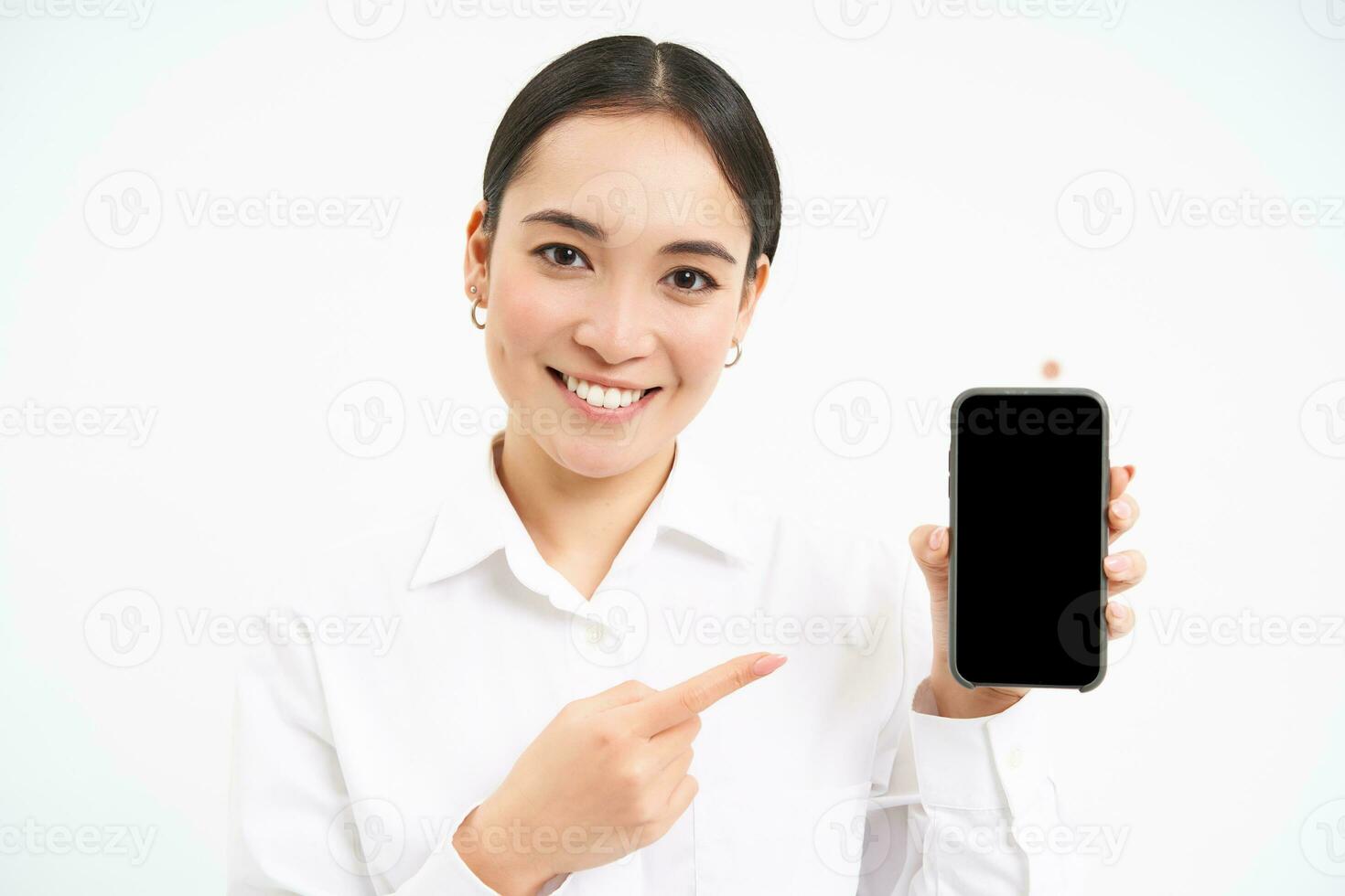 Portrait of businesswoman showing smartphone screen, pointing at mobile phone and smiling, recommending application, standing over white background photo
