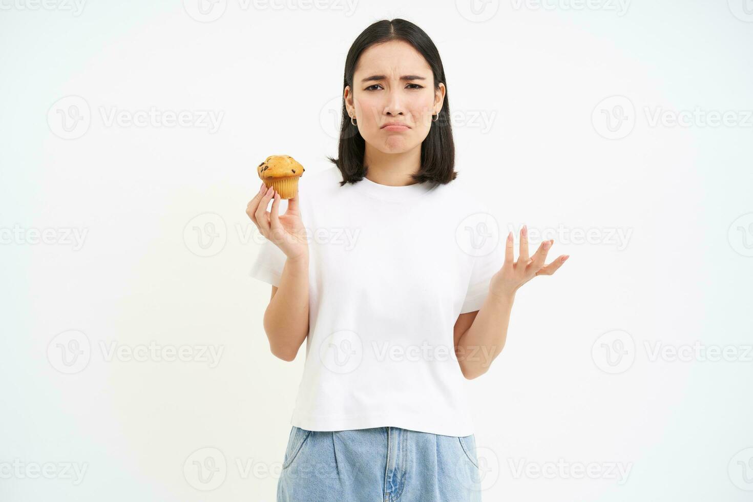 Upset asian woman, holding cupcake and complains, wants to eat junk sweet food, stands over white background photo