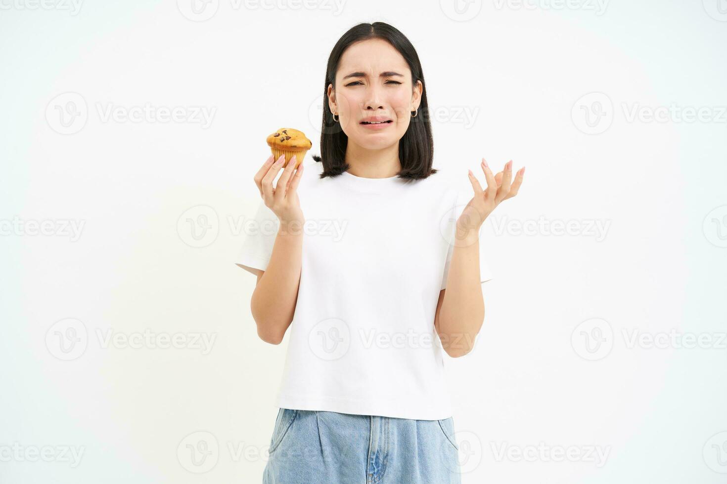 retrato de infeliz coreano mujer mira a magdalena con triste rostro, niña en dieta hipocresía comer Pastelería, concepto de sano alimento, blanco antecedentes foto