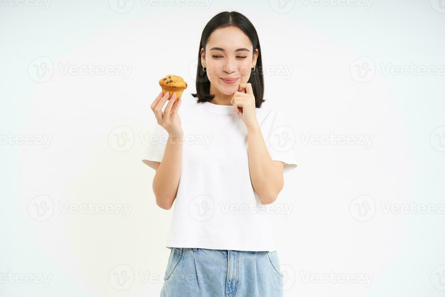 Image of young modern woman with cupcake, looks at delicious pastry from bakery with happy face, white background photo