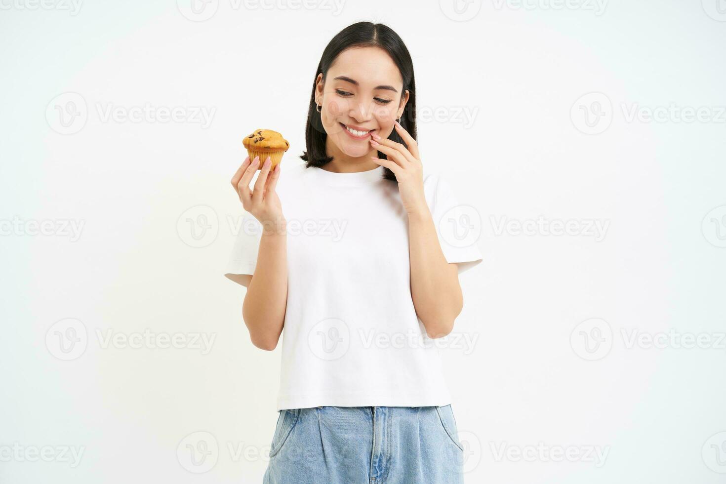 Bakery and sweets. Happy asian woman looking at tasty cupcake, eating pastry, white background photo