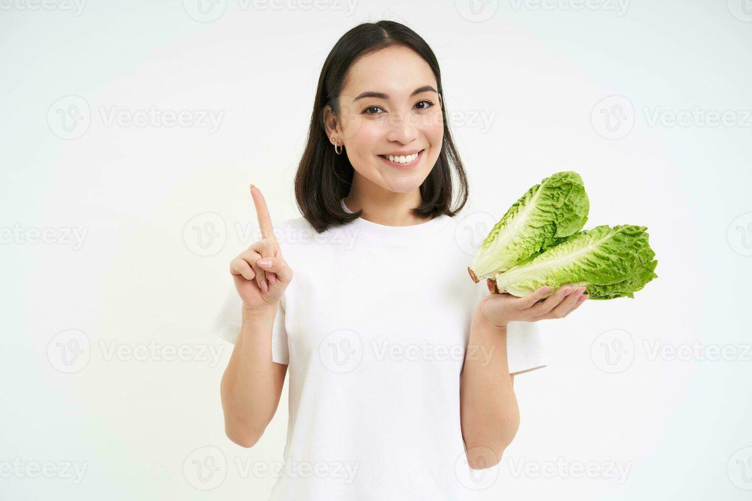 imagen de sonriente asiático niña muestra repollo y uno dedo, puntos, enseña sano dieta, soportes terminado blanco antecedentes con lechuga foto