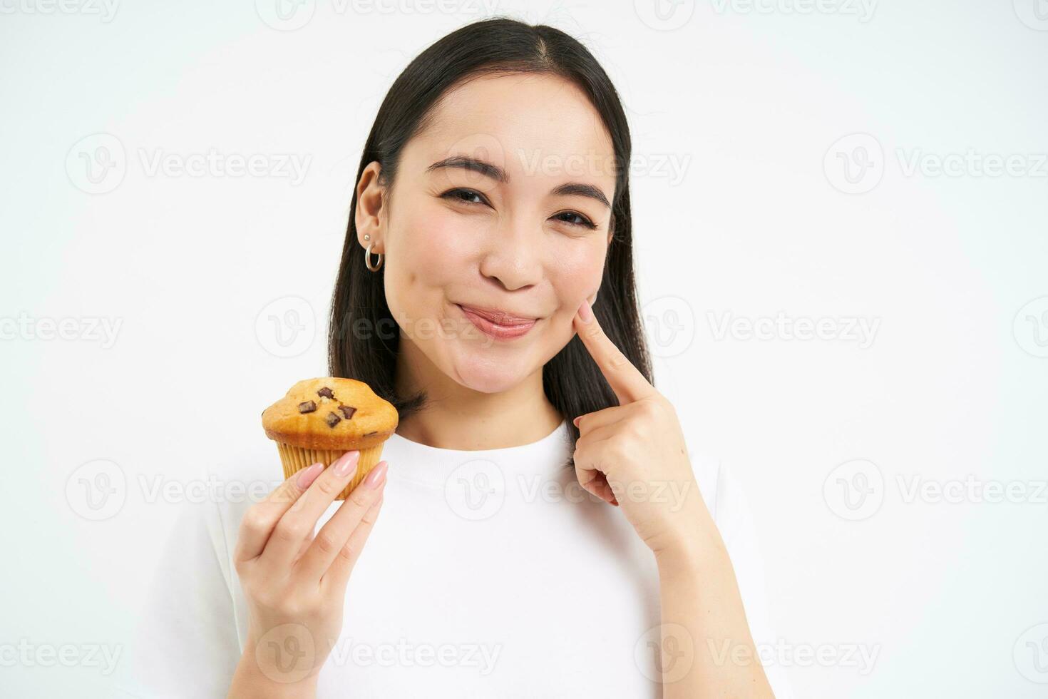 Close up portrait of smiling korean woman, eating cupcake with pleased face, white background photo