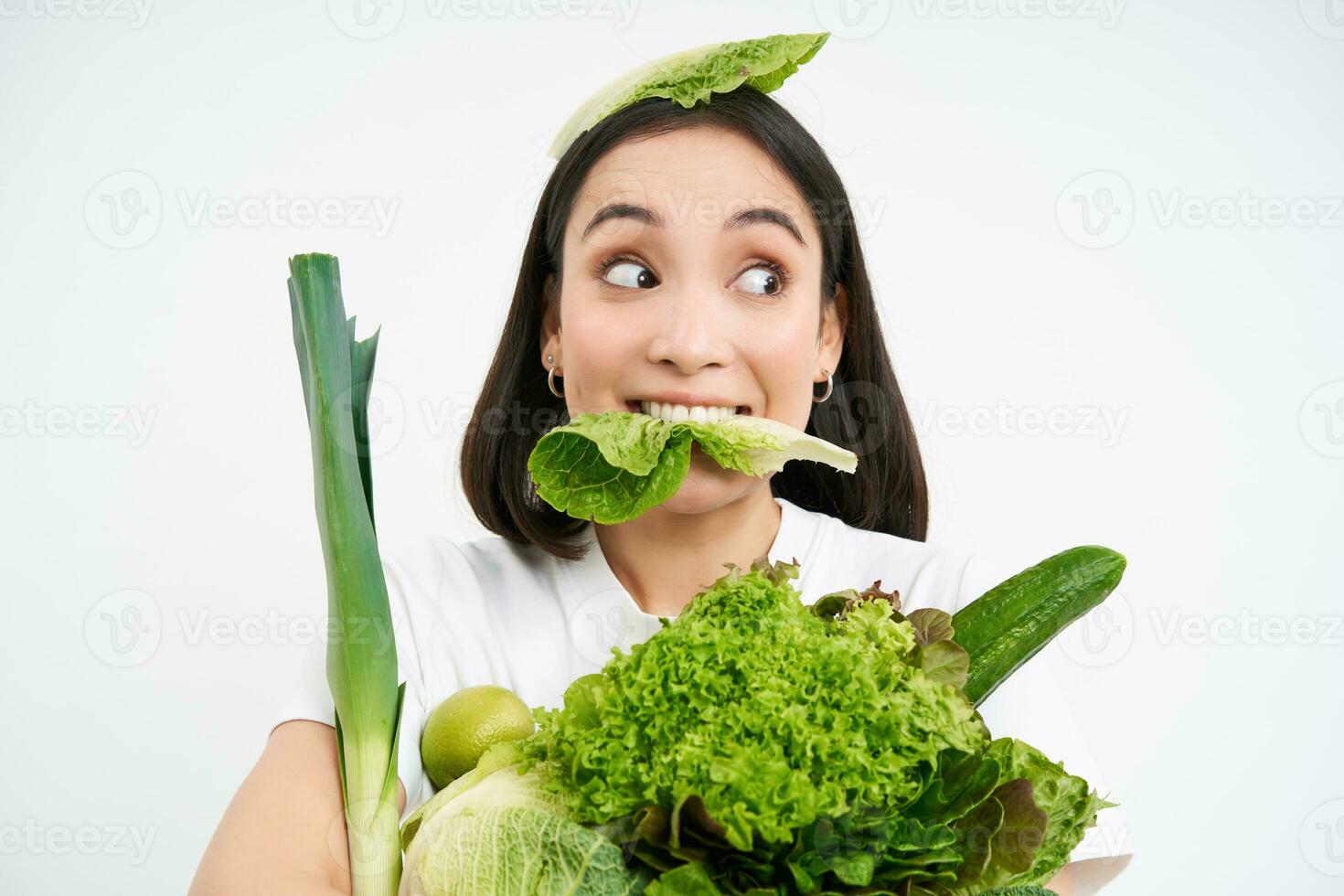 Close up portrait of asian girl munching lettuce leaf, looking excited, holding pile of vegetables, green organic food from garden, standing over white background photo