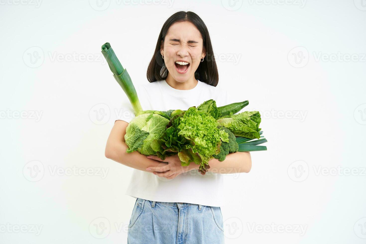 Image of sad, unhappy girl on diet, holding vegetables, green raw food, complaining, standing over white background photo