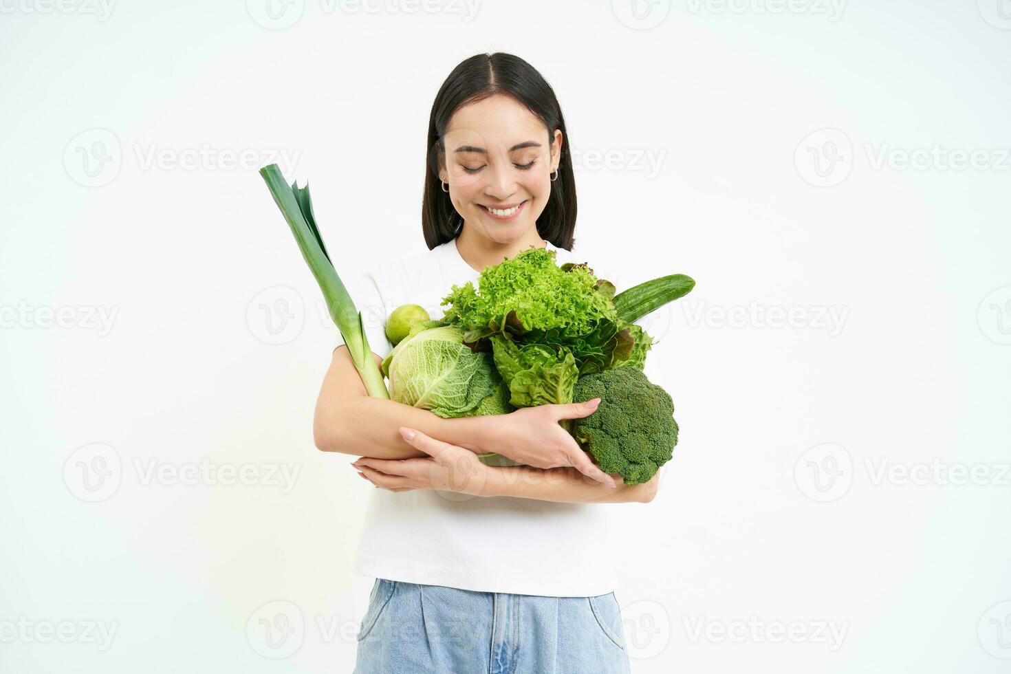 retrato de sonriente coreano mujer, abrazos su verduras, gustos comiendo crudo alimento, vegetariano disfruta nutritivo dieta, blanco antecedentes foto