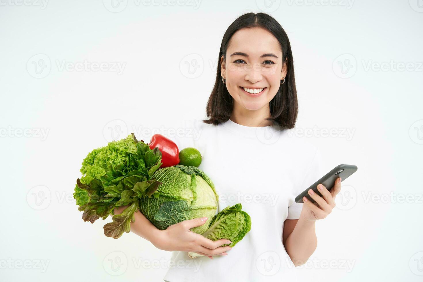 Portrait of happy, smiling korean woman, orders green nutritious food online, holds vegetables and smartphone, standing over white background photo