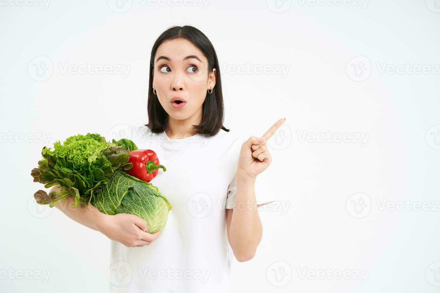 Portrait of girl with surprised face, pointing at advertisement, holding green vegetables, concept of dietry, white background photo