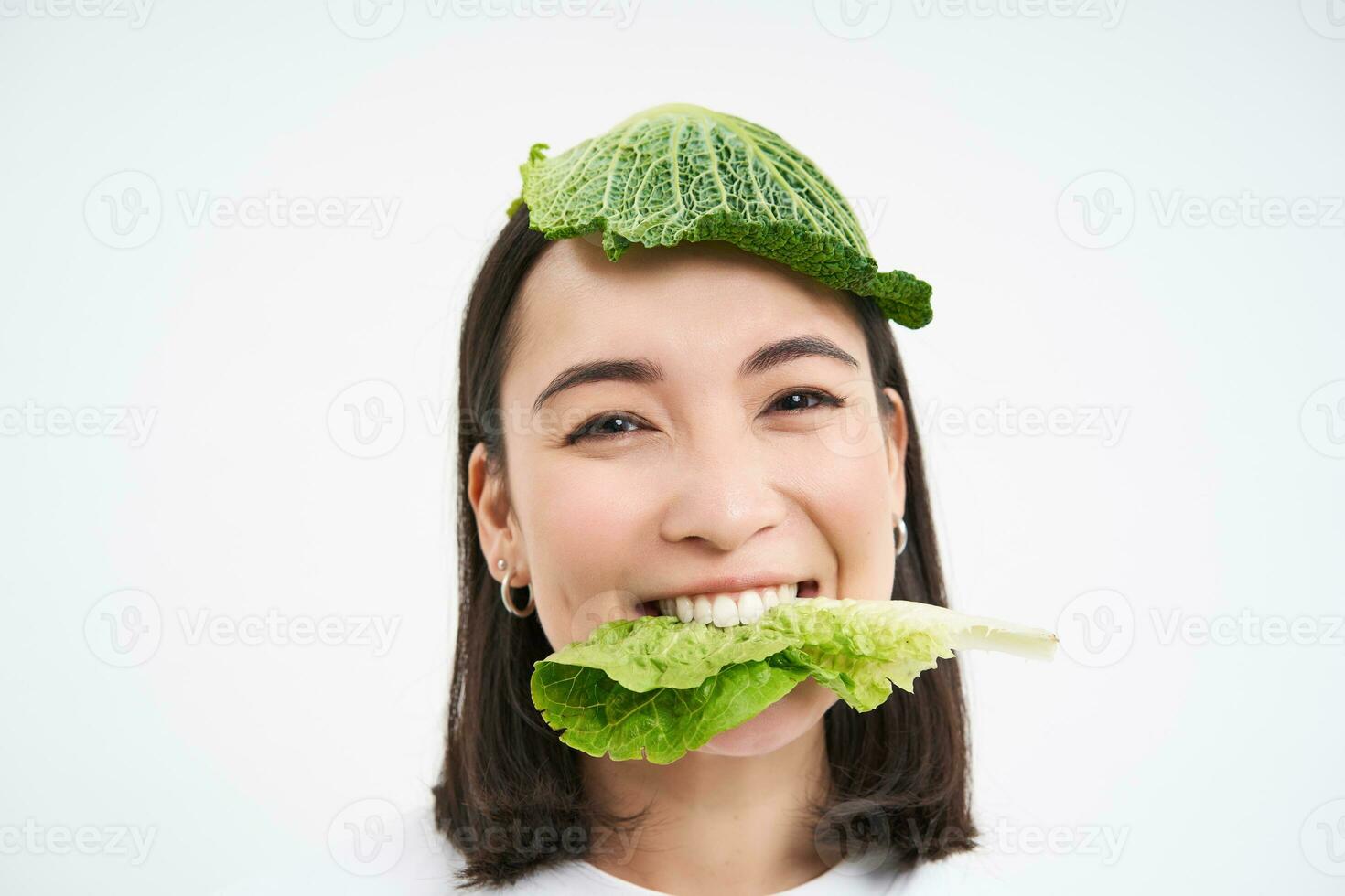 hermosa asiático niña con lechuga en cabeza, sonriente y comiendo repollo hoja, vegano con verduras, blanco antecedentes foto