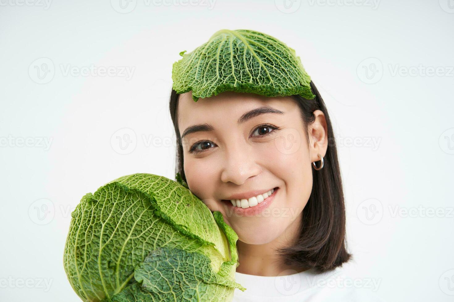 Close up portrait of cute asian woman with lettuce leaf on head, smiling and showing green cabbage, white background photo