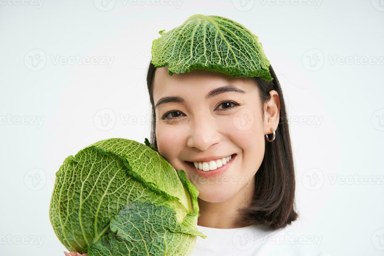 Close up portrait of cute asian woman with lettuce leaf on head, smiling and showing green cabbage, white background photo