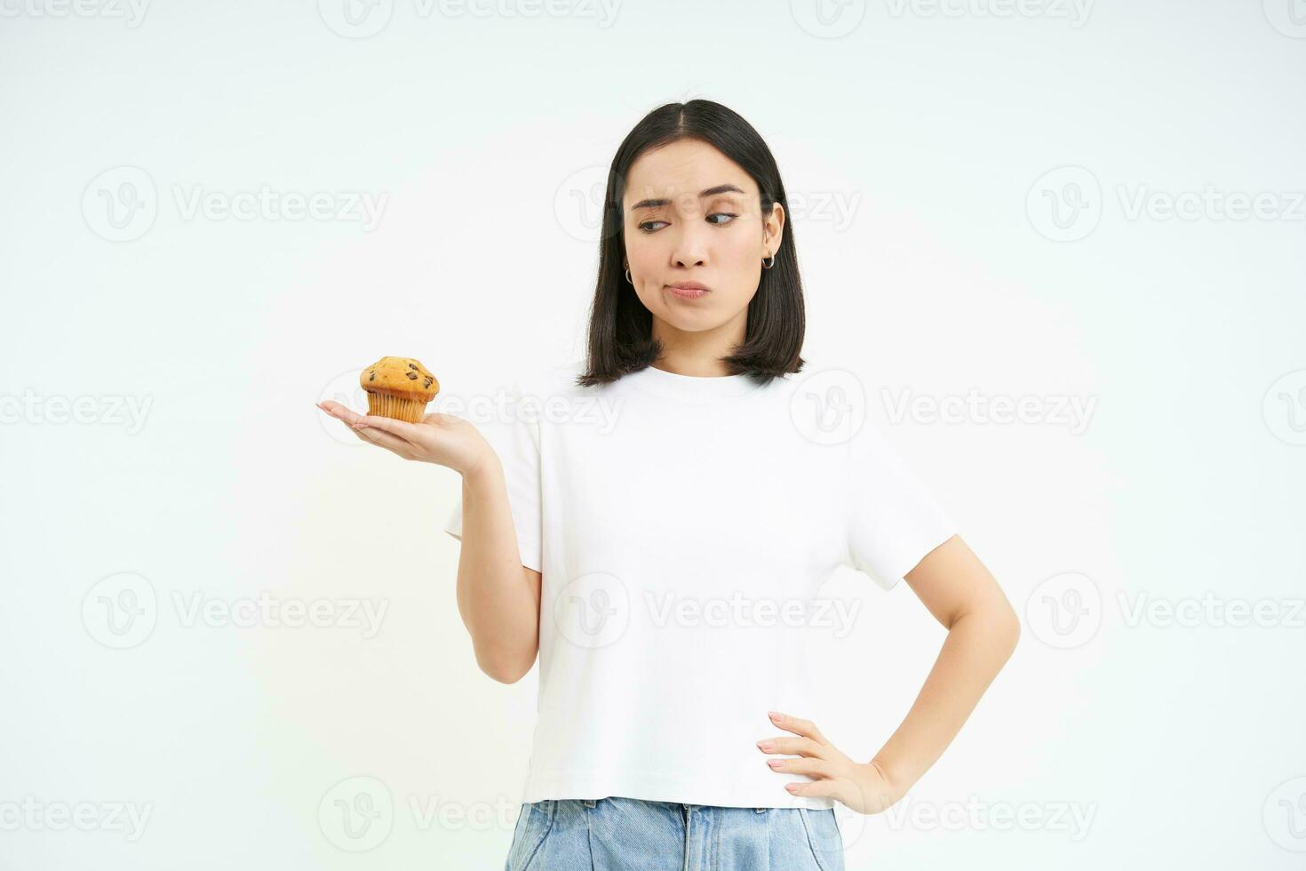 Image of korean girl, looking at cupcake and thinking, making decision, white background photo