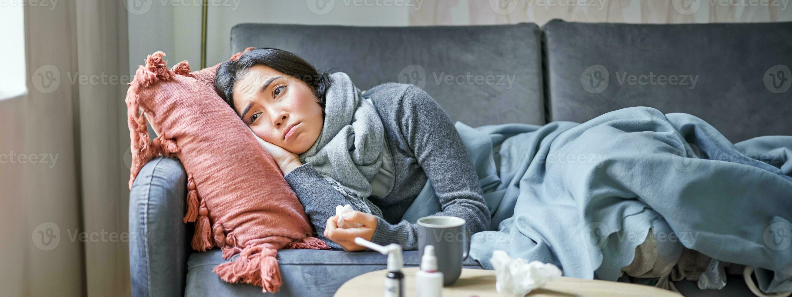 Sick sad korean woman lying on sofa, feeling unwell, catching cold, flu and temperature, looking upset, taking medication photo