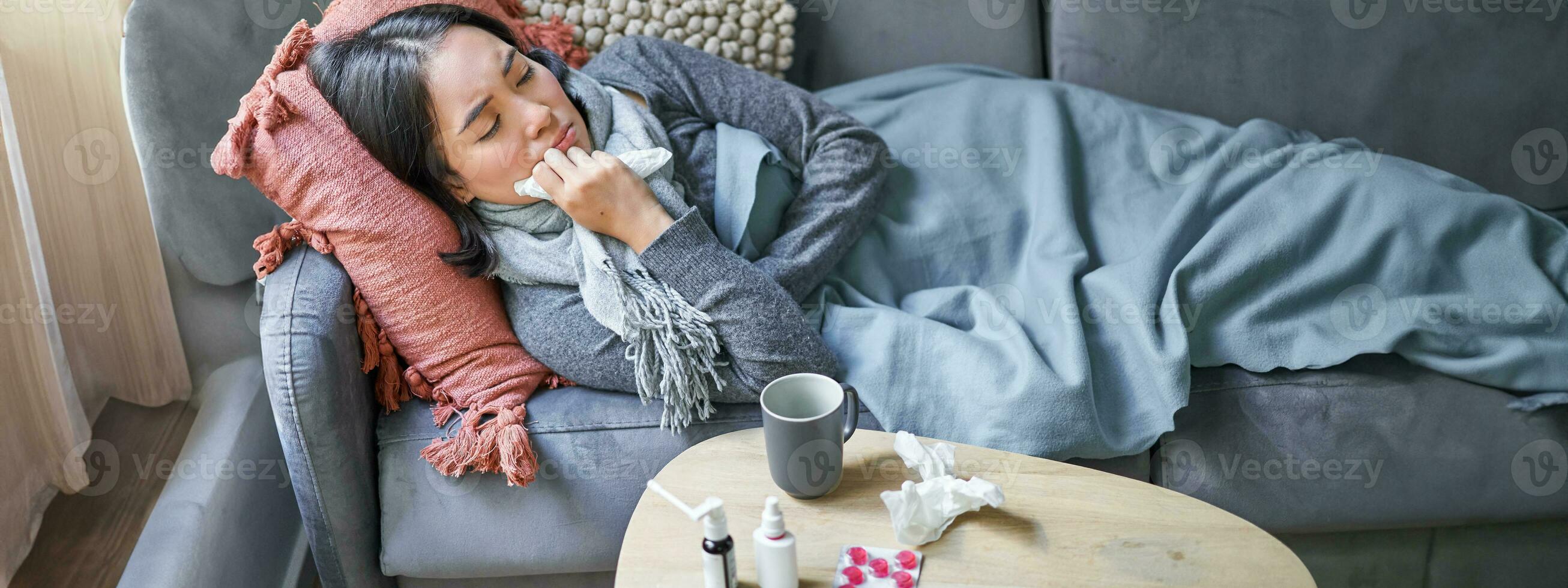 Sick sad korean woman lying on sofa, feeling unwell, catching cold, flu and temperature, looking upset, taking medication photo