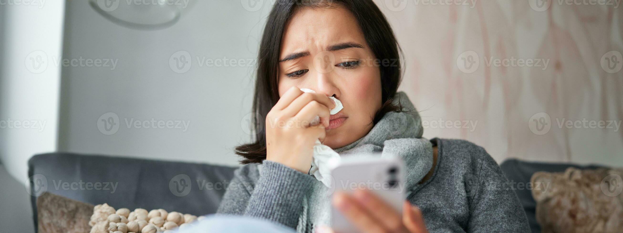 Ill korean woman sitting at home, sneezing, caught cold, reading news on mobile phone, freezing at her home because of heating problems photo