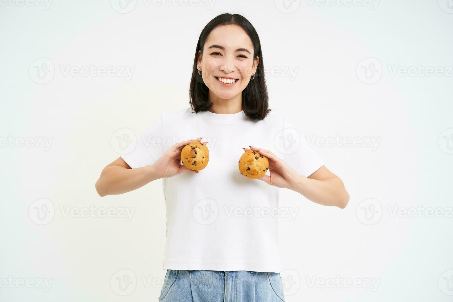 Happy japanese woman shows cupcakes on chest, eating pastry, standing over white background photo