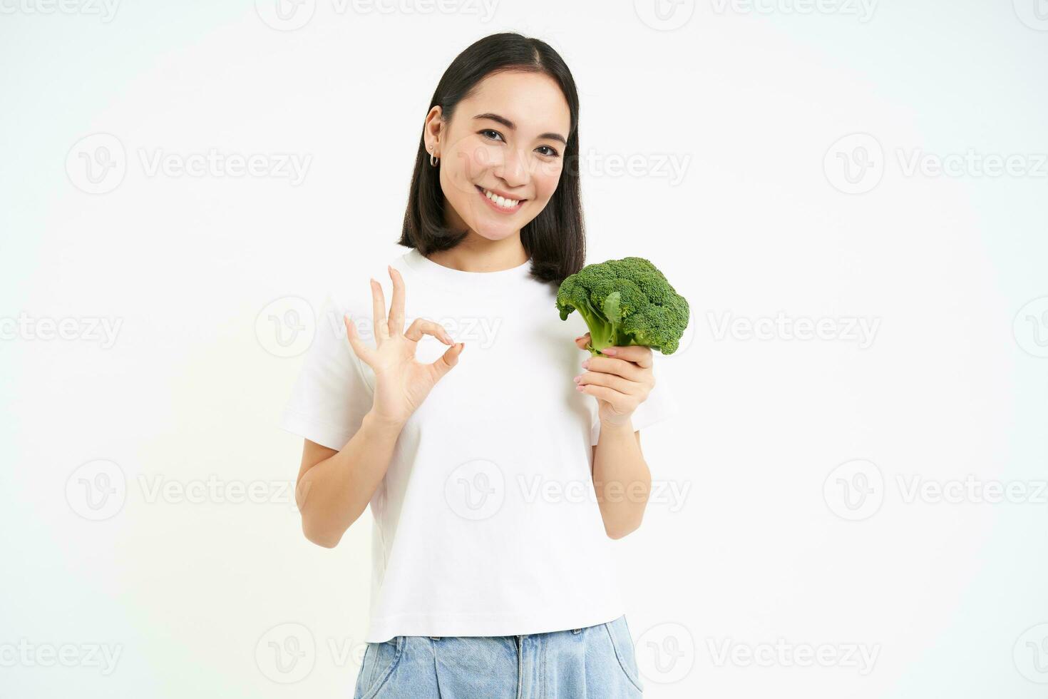 Portrait of healthy smiling woman, shows broccoli and thumbs up, recommends sticking to nutritious diet with vegetables, white background photo