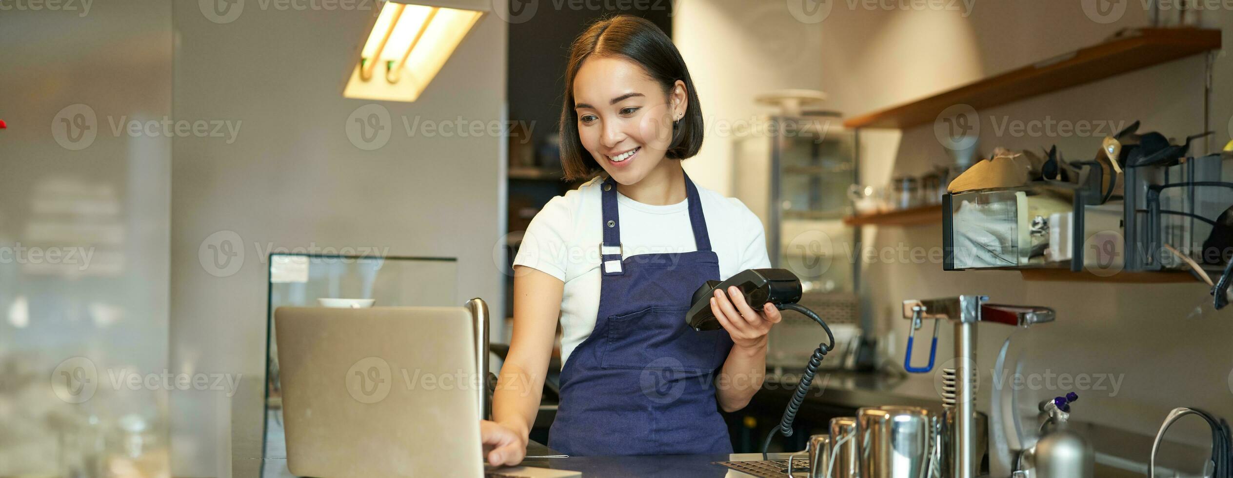 Smiling asian barista, girl with card terminal, payment machine and laptop, standing in cafe, processing payment for coffee order photo