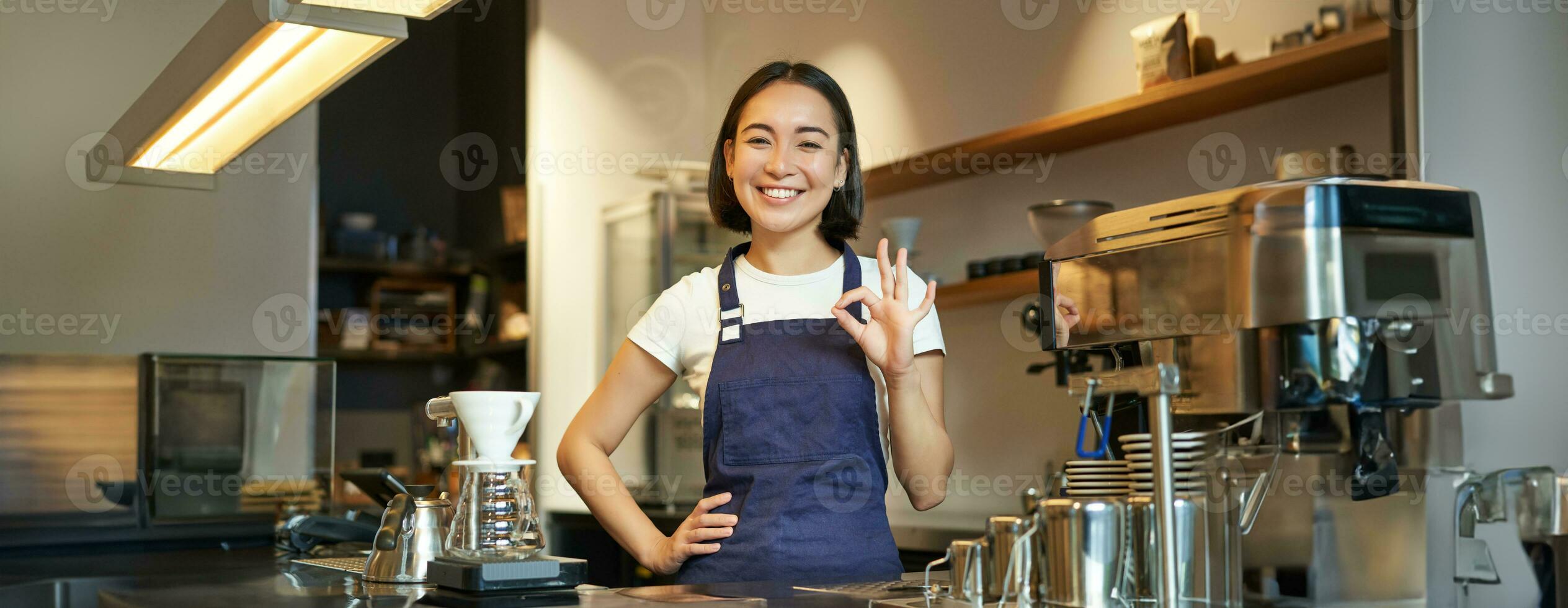 Portrait of smiling korean barista, girl at the counter, wears blue apron, works in coffee shop, shows okay sign photo