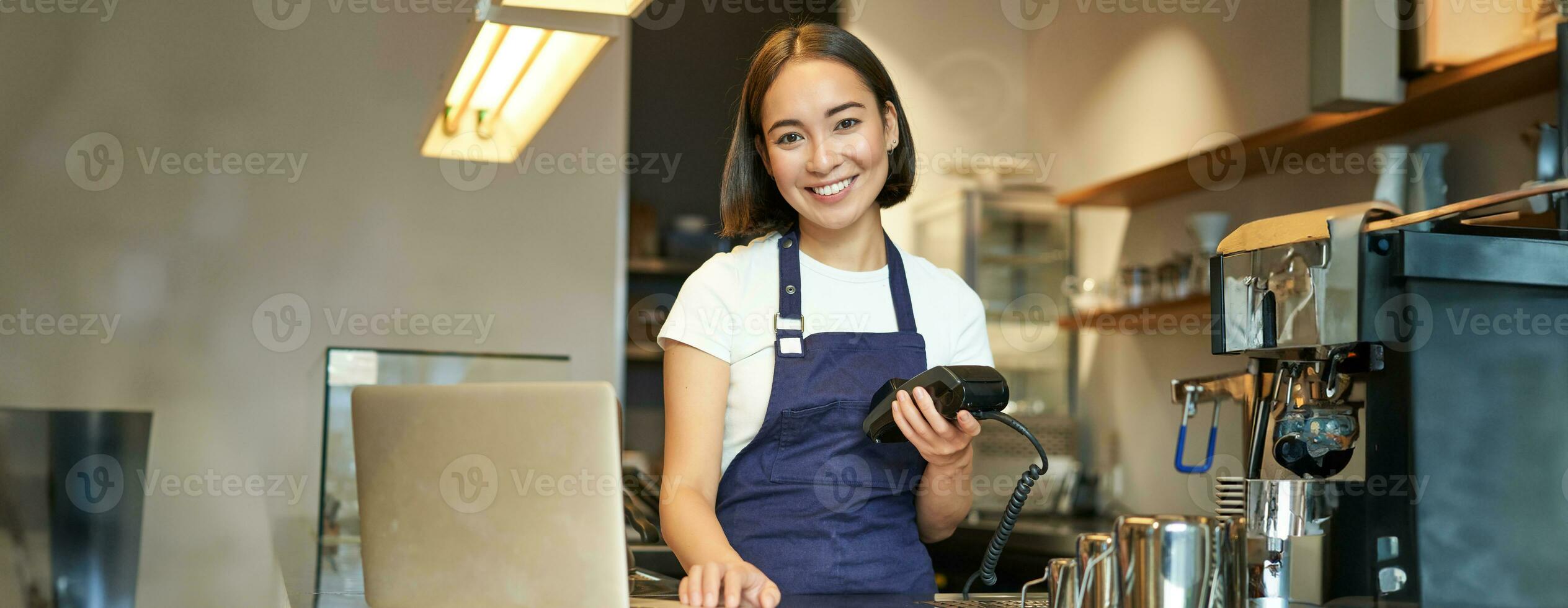 Smiling asian barista, girl with card terminal, payment machine and laptop, standing in cafe, processing payment for coffee order photo